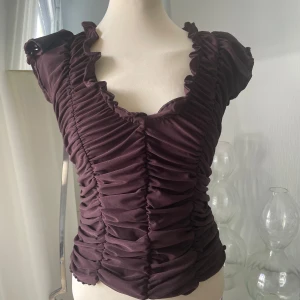 Brun volangblus från Catwalk - Supersnygg brun blus från Catwalk med volangkantad v-ringning och rynkade detaljer framtill. Kortärmad och figurnära passform i mjuk polyesterblandning. Perfekt för dig som vill sticka ut med en cool och trendig topp.