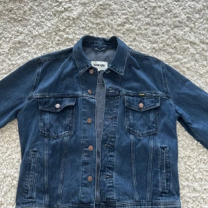 Blå jeansjacka från Wrangler, stl L - Klassisk blå jeansjacka från Wrangler i storlek L. Jackan har två bröstfickor med lock och knappar, samt sidofickor. Snygga kopparfärgade knappar med Wrangler-logga och coola söm-detaljer. Perfekt för dig som gillar tidlös denimstil.