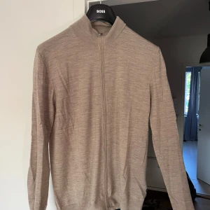 Beige merinoullskofta från Martinique - Säljer en stilren beige kofta från Martinique i 100% merinoull. Koftan har hel dragkedja framtill, hög krage och långärmad passform. Perfekt för lager-på-lager och ger en clean look till din outfit.