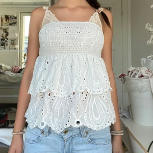 Vit broderad tubtopp - Supersöt vit linnetopp med broderade detaljer. Toppen har smockad överdel och volangig nederdel med hålbroderier och vågig kant. Perfekt för varma dagar och ger en romantisk vibe till din outfit.