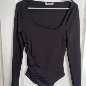 Svart asymmetrisk body från NA-KD - Svart långärmad body från NA-KD med asymmetrisk fyrkantig ringning och tight passform. Toppen har en snygg rynkdetalj längs sidan i stretchigt material. 
