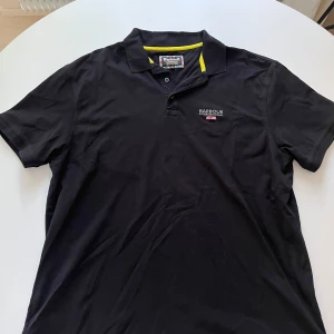 Svart pikétröja från Barbour XXL - Svart pikétröja från Barbour International i bomull med klassisk krage, korta ärmar och broderad logga på bröstet. Snygga gula detaljer på insidan av kragen ger en cool kontrast. Perfekt för dig som gillar stilren och sportig look.