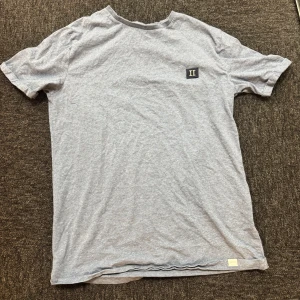 Grå les deux t shirt - Snygg grå t-shirt från Les Deux med liten svartvit logga på bröstet. Klassisk rund hals och korta ärmar. Tillverkad i mjuk bomull som känns skön mot huden. Perfekt för en clean och avslappnad look.