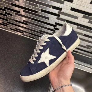 Golden Goose sneakers med stjärna - Snygga sneakers från Golden Goose i mörkblå mocka med vit stjärndetalj på sidan och vit hälkappa. Skorna har vita snören och platt sula, samt en klassisk rund tå. Perfekta för dig som gillar en cool och avslappnad stil.