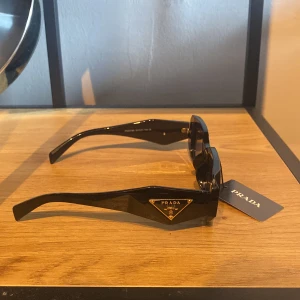 Svarta solglasögon från Prada - Snygga svarta solglasögon från Prada med rektangulära bågar och breda skalmar. Ikonisk guldfärgad Prada-logga på sidan och mörka linser. Tillverkade i Italien och perfekta för en trendig look.