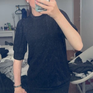 Glittrig svart topp från Vila XS - Snygg svart glittrig topp från Vila i storlek XS. Toppen har halvhög krage, puffiga halvlånga ärmar och en figurnära passform. Perfekt för att sticka ut med lite extra glitter och coola detaljer.