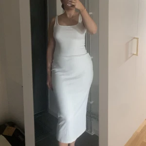 Vit ribbad långklänning med smala axelband - Säljer en stilren vit långklänning med smala axelband och ribbad struktur. Klänningen har en tight passform och är ärmlös, perfekt för varma dagar. Enkel och clean design som är lätt att styla med accessoarer. Jag är 163 och den går nästan ner till fötterna