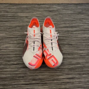 Puma Ultra 5 Ultimate FG - Säljer ett par Puma Ultra fotbollsskor i vitt och neonorange med svarta detaljer. Skorna har snörning, låg profil och mönstrad ovandel i syntetmaterial. Yttersulan har fasta dobbar för bra grepp på planen. Snygg och sportig design med ULTRA-tryck på hälen. De är använda 2 gånger tidigare.