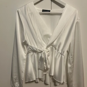 Vit omlottblus med knyt från Missay - Säljer en vit omlottblus från Missay i glansigt material. Blusen har långa ärmar, volang nertill och knyt i midjan för en snygg siluett. Perfekt för dig som gillar feminina detaljer och stilren look.