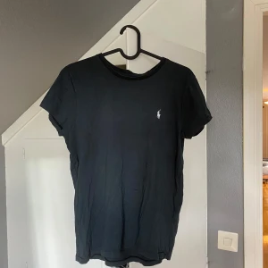Svart t-shirt från Polo Ralph Lauren - Svart t-shirt från Polo Ralph Lauren med klassisk passform och liten vit broderad logga på bröstet. Tillverkad i mjuk bomull och har rund halsringning samt korta ärmar. Perfekt basic-plagg med stilren vibe.