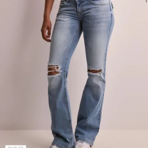 Ljusblå jeans Nelly - Snygga ljusblå bootcut jeans med slitna hål över knäna och klassiska bakfickor med lock och knapp. Jeansen har en normal passform och är tillverkade i mjukt denimtyg. Perfekta för en avslappnad och trendig look. Snörena på knäna har gått upp en del och de är lite slitna nere vid slutet av jeansen pga att jag gått på de