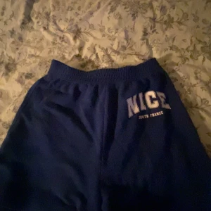 Blå shorts H&M NICE South France - Blåa shorts från H&M med trycket 'NICE South France' på ena benet. De har en elastisk midja och är gjorda i mjukt material, perfekt för lugna/chill dagar.