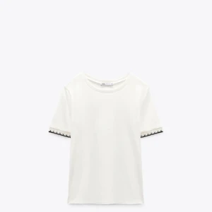 Vit t-shirt med spetsdetalj Zara - Vit t-shirt från Zara med korta ärmar och snygg svart o guldig spetsdetalj och broderi vid ärmsluten.