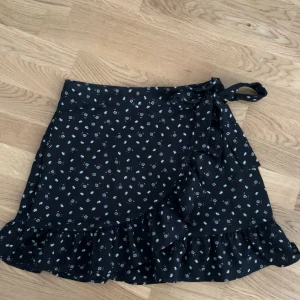 Svart blommig omlottkjol med volang - Supersöt svart kortkjol med små vita blommor, omlottmodell och knyt i midjan. Kjolen har volang längs nederkanten som ger en extra touch. Storlek M men sitter som S!