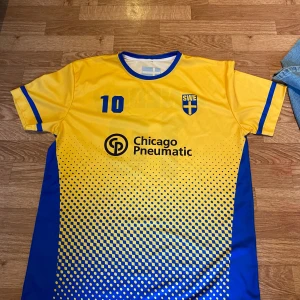 Svensk fotbollströja nummer 10 gul/blå - Gul och blå fotbollströja med nummer 10 och texten SWEDEN på ryggen. Framsidan har SWE-emblem och sponsorlogga. Tröjan har korta ärmar, blå detaljer och ett prickigt mönster nedtill. Tillverkad i lätt och ventilerande material, perfekt för match eller träning.