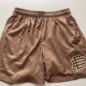 Beige mönstrade shorts - Snygga beige shorts mönster över hela tyget och en vit broderad logga på ena benet. De har elastisk midja för skön passform och är perfekta för sommaren. Materialet känns lyxigt och andas bra.