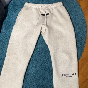 Essentials Fear of God mjukisbyxor ljusgrå - Ljusgrå mjukisbyxor från Essentials Fear of God med elastisk midja och snörning. Byxorna har raka ben, sidofickor och en bakficka. Mjukt bomullsmaterial och logga tryckt på benet. Perfekta för chill eller streetwear.