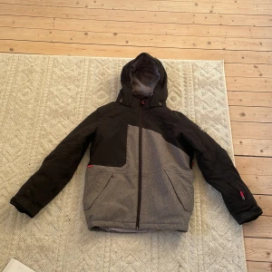 Svart och grå pufferjacka med huva - Snygg pufferjacka i svart och grått med huva och dragkedja framtill. Jackan har två stora fickor och coola röda detaljer vid dragkedjorna. Perfekt för kalla dagar när du vill hålla stilen och samtidigt vara varm.