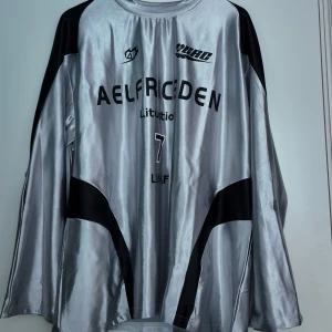 AELFRIC EDEN TRÖJA - Snygg långärmad fotbollströja i blankt silver med svarta detaljer och tryck. Tröjan har rund hals, svarta paneler på sidor och ärmar samt tryck med texten Aelfriceden och nummer 7. Materialet är glansigt och känns sportigt. Tröjan är helt oanvänd och togs endast ut ur dens påse för att ta bilder på den. Pris kan diskuteras. 