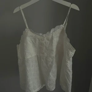Vit broderad linnetopp från H&M - Supersöt vit linnetopp från H&M med broderade blommor och spetskant. Toppen har smala axelband, dekorativa knappar framtill och en luftig passform. Perfekt för varma dagar! Nytt skick! Endast använd en gång.