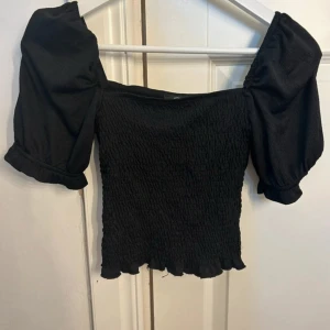 Svart smockad topp med puffärmar - Supersöt svart topp med smockad kropp och stora puffärmar. Toppen har en croppad passform och volangkant nertill. Perfekt för dig som gillar trendiga detaljer och vill sticka ut lite extra.
