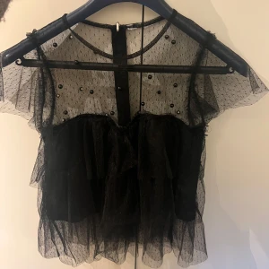 Svart volangblus med pärldetaljer Zara - Supersöt svart blus från Zara i mesh med volanger och pärldetaljer. Blusen har korta ärmar, transparent tyg och dragkedja bak. I strl Xs men klippt av lappen då den syntes ibak🥰