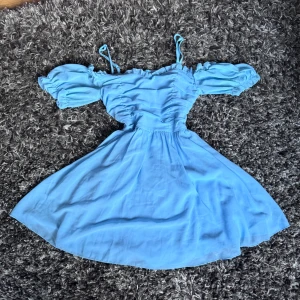 Ljusblå offshoulder klänning Shein XS - Supersöt ljusblå kortklänning från Shein i storlek XS. Klänningen har offshoulder puffärmar, smala axelband och rynkade detaljer över bysten. Kjolen är utsvängd och materialet känns lätt och flowy, perfekt för varma dagar.