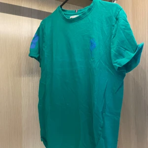 Grön t-shirt från U.S. Polo Assn. - Snygg grön t-shirt från U.S. Polo Assn. med klassisk passform och korta ärmar. T-shirten har en liten broderad logga på bröstet och är tillverkad i mjuk bomull. Perfekt för en chill och avslappnad stil.