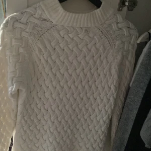 Vit stickad tröja med flätmönster - Säljer en vit stickad tröja med snyggt flätmönster och hög ribbad krage. Perfekt för kyliga dagar och ger en mysig vibe. Tröjan har långärmade ärmar och är riktigt mjuk och skön att ha på sig.