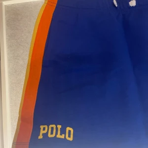 Blå kjol Polo Ralph Lauren S - Blå kjol från Polo Ralph Lauren med gula och orangea ränder längs sidorna. Klassisk POLO-logga i gult på ena benet. Perfekta för sommaren och speciellt när man spelar golf. Elastisk midja och skön passform. Stl barn XL vilket motsvarar ca en S