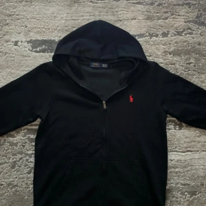 Polo Ralph Lauren kofta - Svart hoodie med dragkedja från Polo Ralph Lauren. Klassisk design med huva, två fickor framtill och röd broderad logga på bröstet. Perfekt för en avslappnad stil.