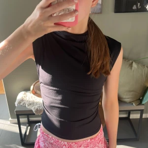 Svart ärmlös topp med rynkade sidor - Säljer en svart, tight och ärmlös topp med hög hals och snygga rynkade detaljer på sidorna. Toppen är croppad och gjord i ett stretchigt, mjukt syntetmaterial som sitter riktigt snyggt på kroppen. Perfekt till jeans eller kjol för en clean look.