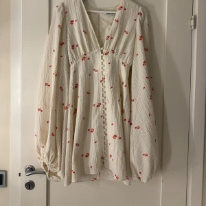 Blommig vit klänning från Adoore - Supersöt vit kortklänning med små röda blommor från Adoore. Klänningen har v-ringning, långa puffärmar och knappar framtill. Använd fåtal gånger, inga defekter. 💞