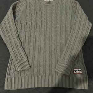 Beige kabelstickad tröja  - Kabelstickad beige tröja från Iconic Sport med rund halsringning och långa ärmar. Tröjan har breda ribbade muddar vid ärmslut och nederkant samt en snygg patch nertill. Perfekt för dig som gillar klassisk och stilren look.