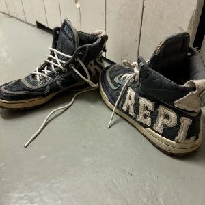 Replay höga sneakers med broderi - Unika höga sneakers från Replay i mörkblått tyg med vita detaljer och stora broderade bokstäver på sidorna. Skorna har vit snörning, beige sula och vadderad insida för extra komfort. Perfekta för dig som vill sticka ut med en cool streetstil. Går inte att få tag på idag! 