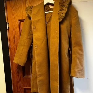 Brun kappa med fuskpäls från H&M - Snygg brun kappa från H&M i storlek XS med huva och fuskpäls runt kanten. Kappan har knappar framtill och ett bälte i midjan för extra stilpoäng. Perfekt för kyliga dagar när du vill vara både varm och trendig.