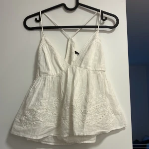 Vit broderad linnetopp från Zara - Superfin vit linnetopp från Zara med tunna axelband som korsas i ryggen. Toppen har v-ringning, broderade detaljer nertill och spetskant längs urringningen. Luftig och flowy passform, perfekt för varma dagar.
