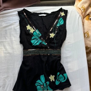 Svart blommig topp från Rocha - Svart mönstrad topp från Rocha John Rocha med gröna och gula blommor, paljettdetaljer och omlott-v-ringning. Toppen är ärmlös och har markerad midja med dekorativa band och paljetter. Perfekt för dig som gillar unika detaljer och färgglada broderier. Jag skulle säga att toppen är lite mer åt det större hållet och är perfekt för sommaren med tanke på dess luftiga tyg. Toppen har paljett och glaspärle detaljer 