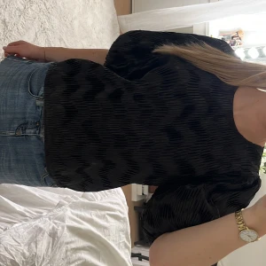 Svart plisserad topp från Neo Noir - Plisserad svart topp från Neo Noir med puffiga korta ärmar och rund halsringning. Toppens tyg har en glansig finish och ett vågigt strukturmönster som ger en cool look. Stängs med knapp i nacken. Perfekt att styla med jeans eller kjol. Hittar inte vilken storlek den är men borde passa från xs-M