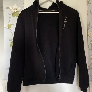Svart fleecejacka från Sail Racing - Svart fleecejacka från Sail Racing med huva och dragkedja hela vägen. Jackan har broderad logga på bröstet och en enkel, clean look. Perfekt för kyliga dagar när du vill vara både bekväm och snygg. Mjuk insida och normal passform.