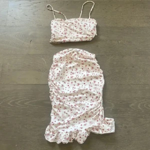 Vit blommig kortklänning Pink & Pink - Supersöt vit kortklänning från Pink & Pink med röda små blommor och tunna axelband. Klänningen har volangkant nertill och är figurnära med rynkade detaljer på sidorna. Perfekt för sommarens alla tillfällen!