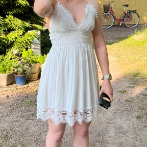 Vit spetsklänning med smala axelband - Superfin vit kortklänning med spetsdetaljer och smala axelband. Klänningen har v-ringad omlott-topp, markerad midja och volangkant nertill. Perfekt för sommarens alla tillfällen och ger en romantisk vibe.