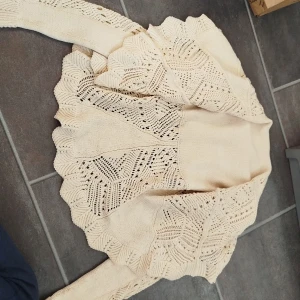 Beige stickad kofta med volangkant - Supersöt beige stickad kofta med volangkant och vackert hålmönster. Koftan har lång ärm och en öppen, svepande passform som ger en romantisk vibe. Perfekt att slänga över en topp för en extra touch till outfiten.