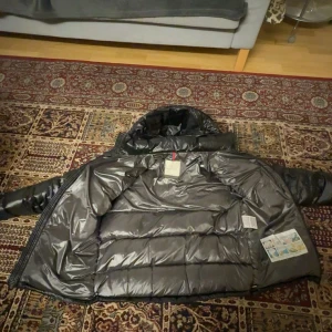 Svart Moncler pufferjacka med huva - Säljer en svart pufferjacka från Moncler med glansig finish, huva och dragkedja framtill. Jackan har Moncler-logga på ärmen, två fickor med dragkedja och quiltad design. Perfekt för kalla dagar och riktigt snygg streetstil.