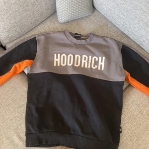 Hoodrich sweatshirt med färgblock - Snygg sweatshirt från Hoodrich med grå och svart färgblock samt orange detaljer längs ärmarna. Stor HOODRICH-logga framtill och HOOD-TECH-tryck på ryggen. Crewneck och långärmad modell i mjuk bomull, perfekt för en streetwear-look.