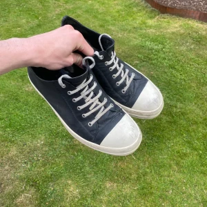 Rick Owens Low Leather Sneakers - Säljer ett par svarta sneakers från Rick Owens med vit tå och sula. Skorna har snörning med vita skosnören och är tillverkade i skinn. Klassisk rund tå och platt sula ger en clean och edgy vibe. Perfekta för dig som gillar streetwear och designerstil. Nypris 8400, slitet men bra skick! Riktigt svåra att få tag på