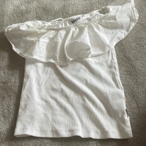 Vit offshoulder topp från Molo - Supersöt vit offshoulder topp från Molo med bred volang runt axlarna. Toppen är ribbad i tyget och har en luftig känsla, perfekt för varma dagar. Snygg och enkel att matcha med både jeans och kjol.