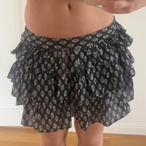 Svart lågmidjad volangkjol med mönster, strl 34 - Supersöt svart voland kortkjol med vita blommiga mönster som har inbyggda shorts. Kjolen har bred resår i midjan där bak men inte fram vilket gör att den sitter extra bra och är tillverkad i ett lätt och luftigt material, perfekt för varmare dagar. Helt ny med prislappen kvar!