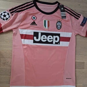 Juventus Pogba 10 rosa matchtröja Adidas - Juventus fotbollströja i rosa med svarta detaljer, Pogba 10 på ryggen. Adidas Climalite-material, kortärmad med svarta ränder på axlarna och Jeep-logga på bröstet. Juventus klubbmärke, mästerskapsmärken och Champions League-patch på ärmen.