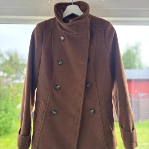 Brun skepparkavaj från H&M, stl 34 - Snygg brun skepparkavaj från H&M i klassisk modell med dubbelknäppning och hög krage. Jackan har långa ärmar och markerad midja för en stilren siluett. Perfekt för dig som vill ha en tidlös look med lite edge.
